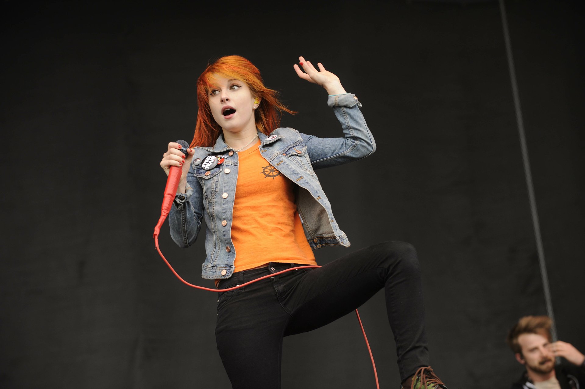 4K Ultra HD PC wallpaper: music artist with orange hair singing into a red mic, wearing denim jacket and orange top, captured mid-performance on stage.