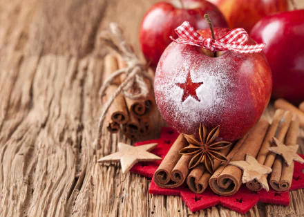 Close-up of a cinnamon-spiced apple decorated with powdered sugar and a red ribbon, surrounded by cinnamon sticks, star anise, and wooden stars on a rustic wooden surface.