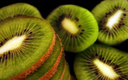 HD PC desktop wallpaper of sliced kiwi fruit close-up: bright green flesh, black seeds and fuzzy brown skin in overlapping slices on a dark background.