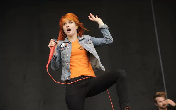 4K Ultra HD PC wallpaper: music artist with orange hair singing into a red mic, wearing denim jacket and orange top, captured mid-performance on stage.