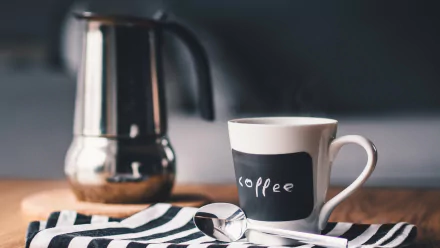 Black and white 4K Ultra HD image of a coffee cup with a spoon and a metallic coffee maker in the background, set on a striped cloth.