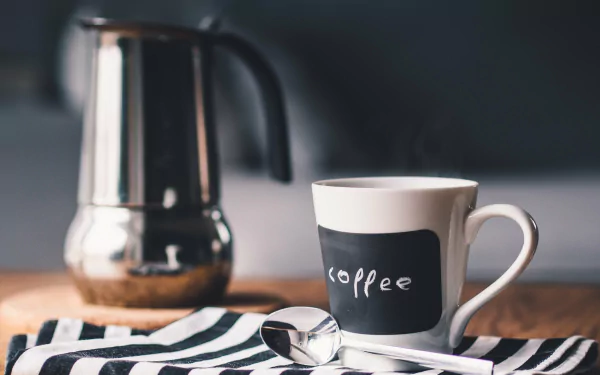 Black and white 4K Ultra HD image of a coffee cup with a spoon and a metallic coffee maker in the background, set on a striped cloth.