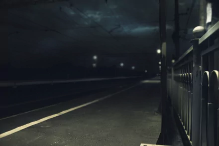 A dimly lit street scene with a railing, captured in 4K Ultra HD. The atmosphere is moody, featuring distant streetlights against a dark sky. Ideal as a desktop wallpaper.