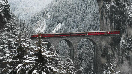 A stunning HD wallpaper featuring a red RhB train crossing an arch bridge amidst a snowy landscape and towering trees, creating a breathtaking winter scene.