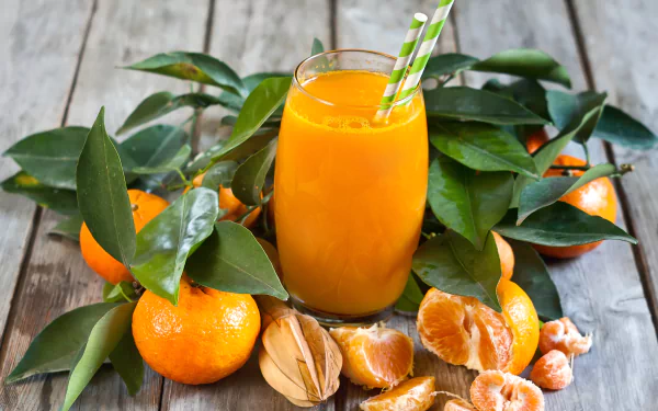 A vibrant HD desktop wallpaper featuring a glass of fresh mandarin juice surrounded by whole and peeled mandarins with green leaves on a rustic wooden surface.