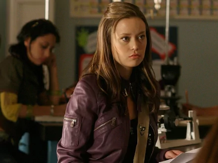 A character from the TV show Terminator: The Sarah Connor Chronicles, depicted in a classroom setting, wearing a purple jacket with a serious expression. HD wallpaper background.