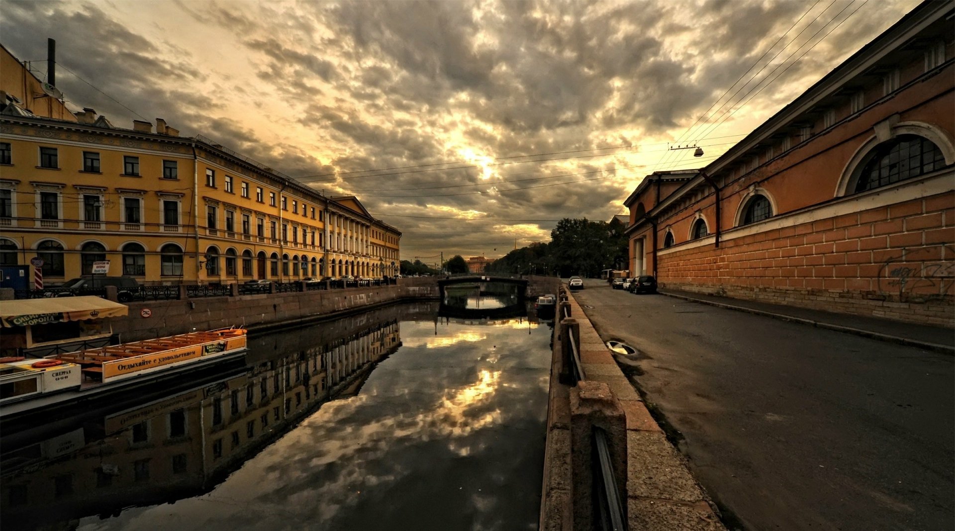 Sunset over a river in Saint Petersburg, Russia, with city buildings reflecting on the water, captured in a dramatic cloudy sky for an HD desktop wallpaper.