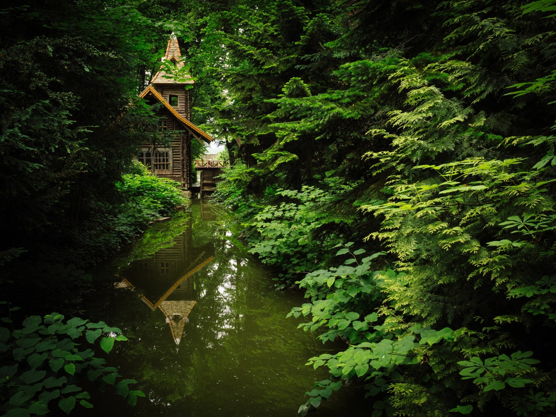 4K Ultra HD PC desktop wallpaper of a man-made watermill cottage reflected in a tranquil forest stream, surrounded by lush green foliage.