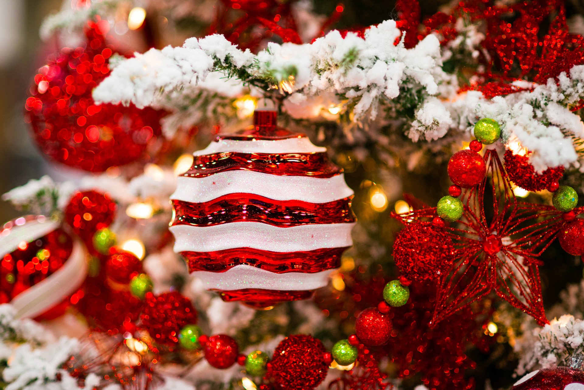 A close-up of red and white striped Christmas ornament hanging on a snowy tree, surrounded by vibrant holiday decorations, creating a festive atmosphere. HD desktop wallpaper.