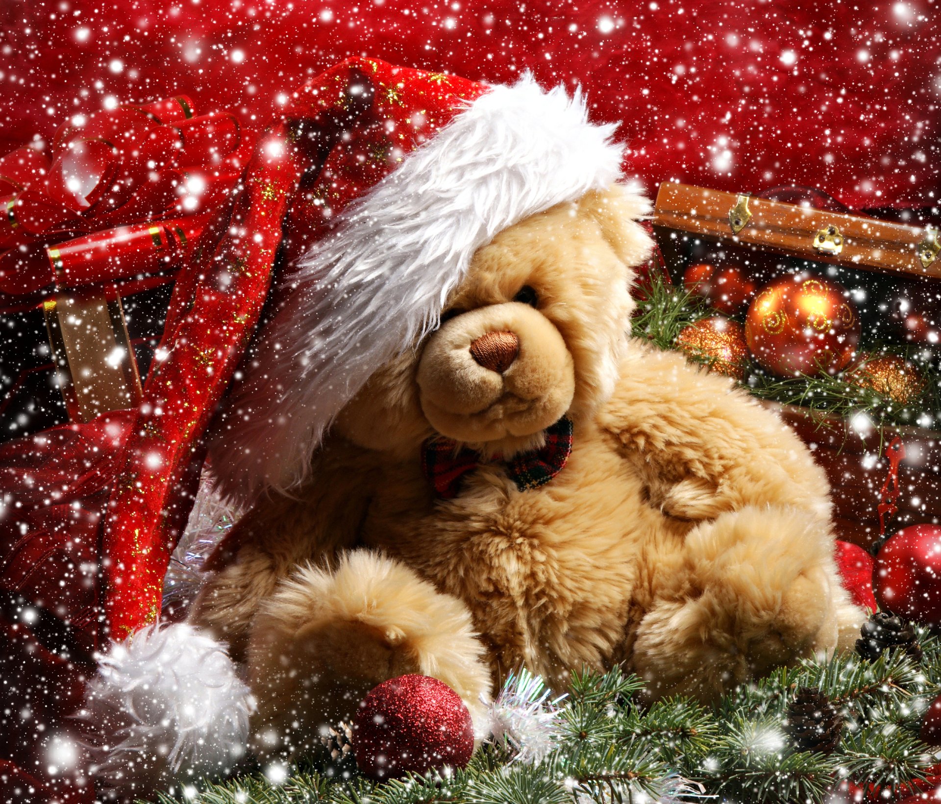 A cozy teddy bear wearing a Santa hat sits among Christmas ornaments and holiday decorations, with a festive snowfall backdrop. HD desktop wallpaper and background.