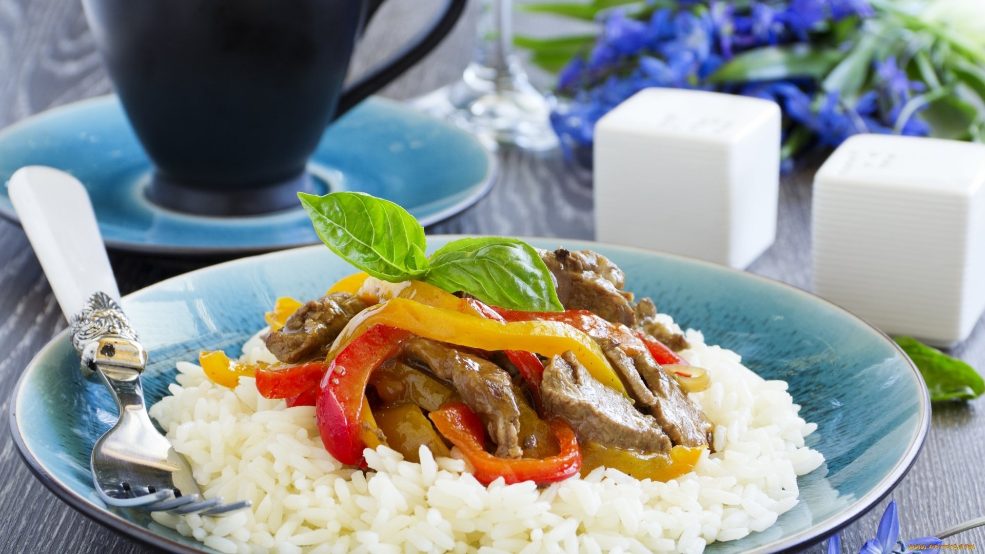 A vibrant meal featuring sliced pepper strips and beef served over a bed of white rice on a blue plate, set against a blurred background with salt shakers and flowers.