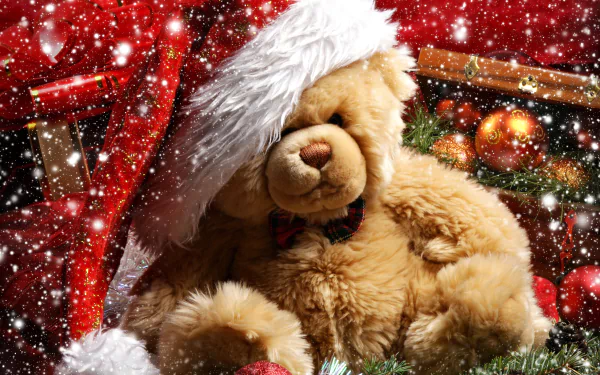 A cozy teddy bear wearing a Santa hat sits among Christmas ornaments and holiday decorations, with a festive snowfall backdrop. HD desktop wallpaper and background.