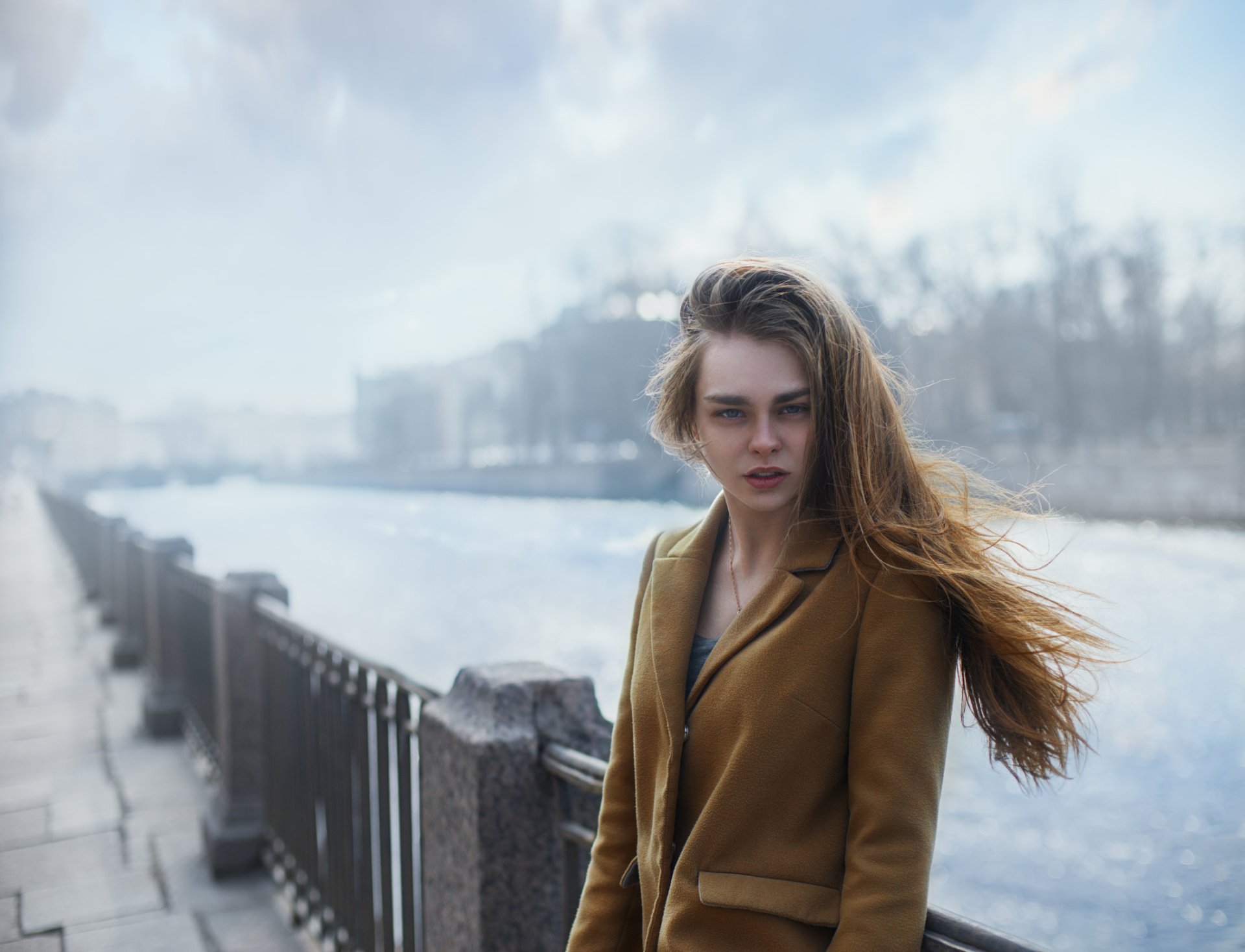 HD desktop wallpaper of a blonde model with long hair and blue eyes standing outdoors by a riverside, wind-tousled hair against a soft bokeh background.