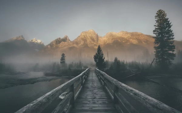 HD wallpaper showing a tranquil landscape with a wooden bridge over a river, leading towards misty mountains surrounded by trees and fog, under a serene sky.
