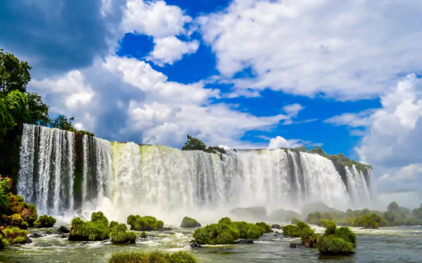 Iguazu Falls cascading under a partly cloudy sky, surrounded by lush greenery and flowing water, captured in an HD desktop wallpaper featuring nature's beauty.
