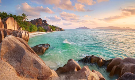 HD PC desktop wallpaper of a tropical Seychelles beach featuring rocky coastline, lush greenery, and calm turquoise waters under a colorful sunset sky.