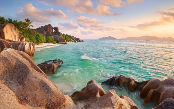 HD PC desktop wallpaper of a tropical Seychelles beach featuring rocky coastline, lush greenery, and calm turquoise waters under a colorful sunset sky.