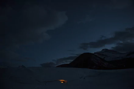 Night scene from The Revenant: snowy plain and shadowed mountains under a dark sky, a lone campfire glow — 2K Quad HD PC desktop wallpaper/background.