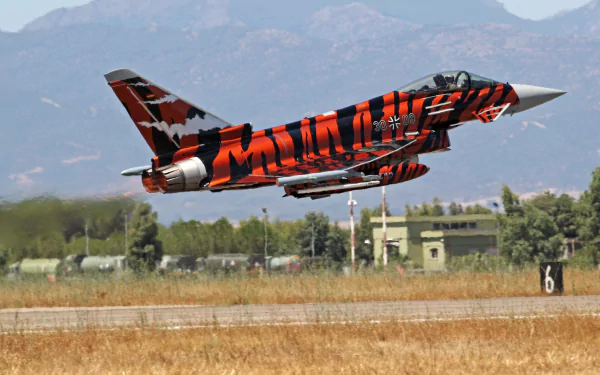 Eurofighter Typhoon warplane with striking orange and black camouflage takes off from a runway, set against a mountainous backdrop, captured in HD for a desktop wallpaper.