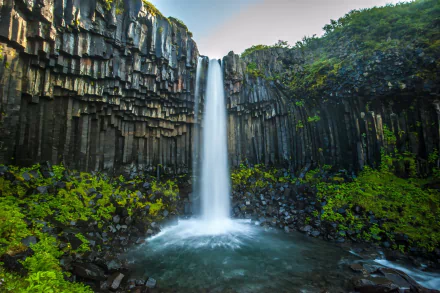2K Quad HD PC desktop wallpaper and background: a slate-column canyon waterfall plunging into a misty pool, framed by vivid mossy greenery under a pale sky.
