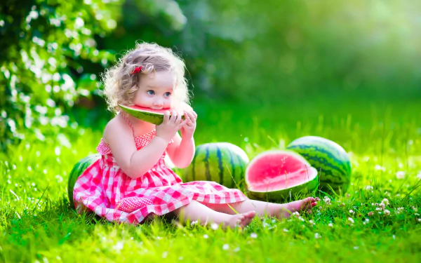 A little girl in a sunny, grassy field enjoys a slice of watermelon. Surrounded by watermelons, she sits barefoot, capturing the essence of summer in vibrant bokeh light.