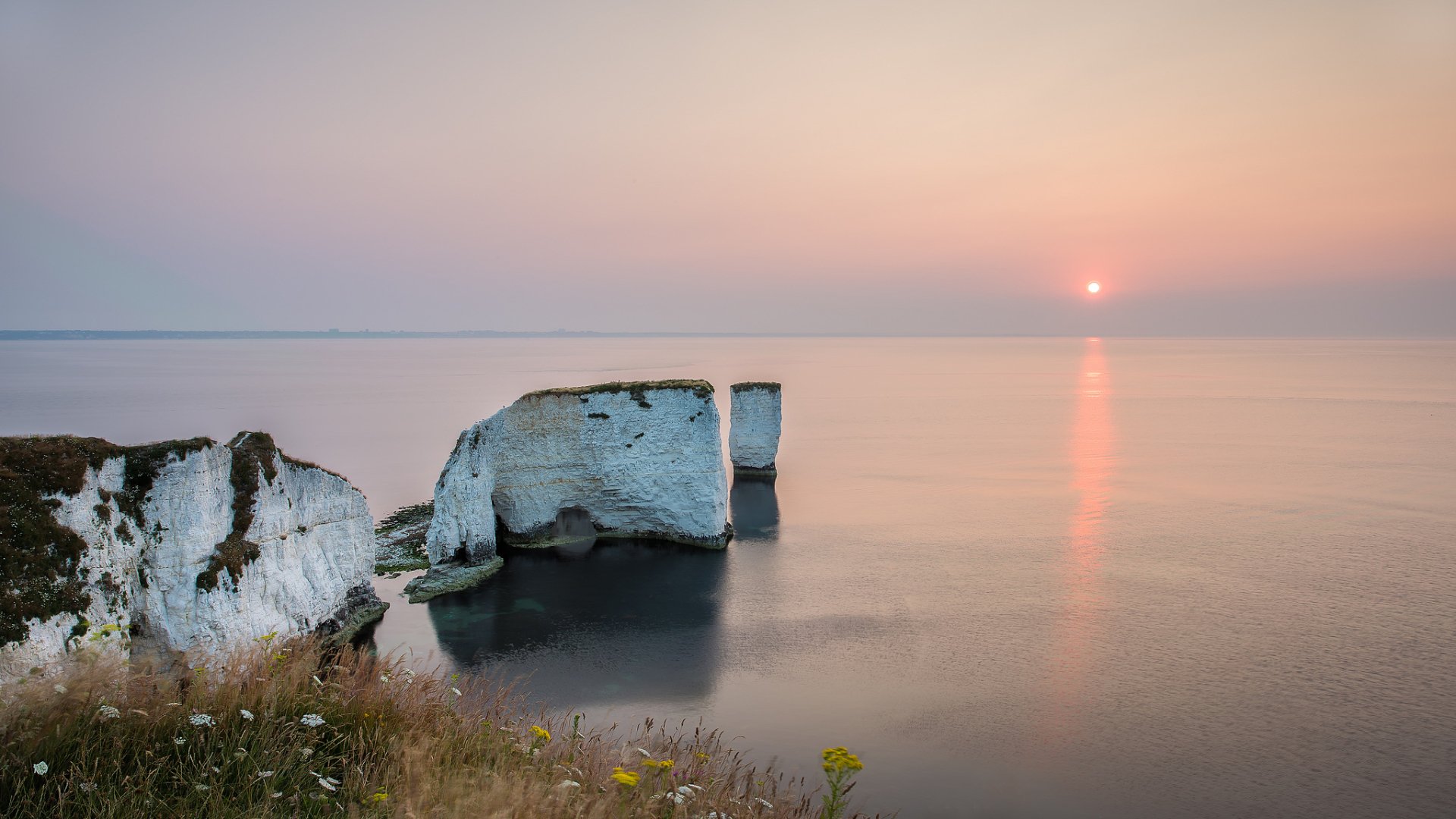 HD desktop wallpaper showing a serene ocean at sunset with cliffs in the foreground and a soft horizon blending sky and water in natural tones.