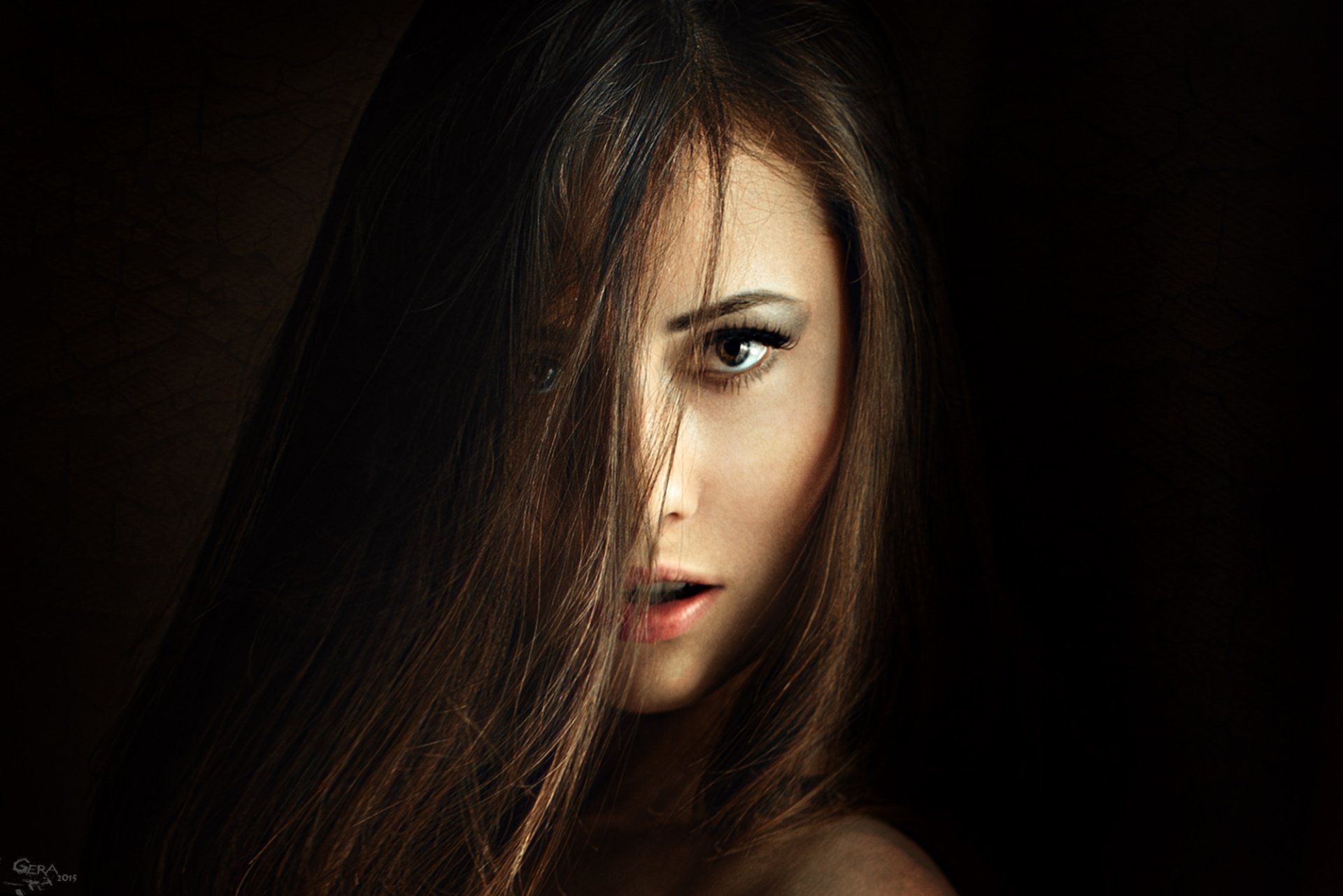 A striking HD desktop wallpaper featuring a brunette woman with brown eyes, her long hair partially covering her face, creating a captivating and mysterious aura.