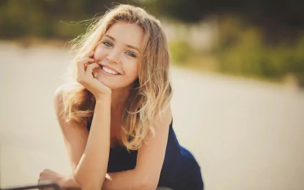 A blonde woman smiles warmly, her hand resting on her cheek, with a bokeh background creating a soft, blurred effect. This HD wallpaper captures her joyful expression in a serene outdoor setting.