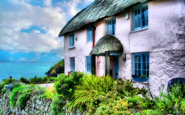 HD desktop wallpaper of a charming man-made cottage in Cornwall, England, featuring a thatched roof and vibrant greenery under a partly cloudy sky.