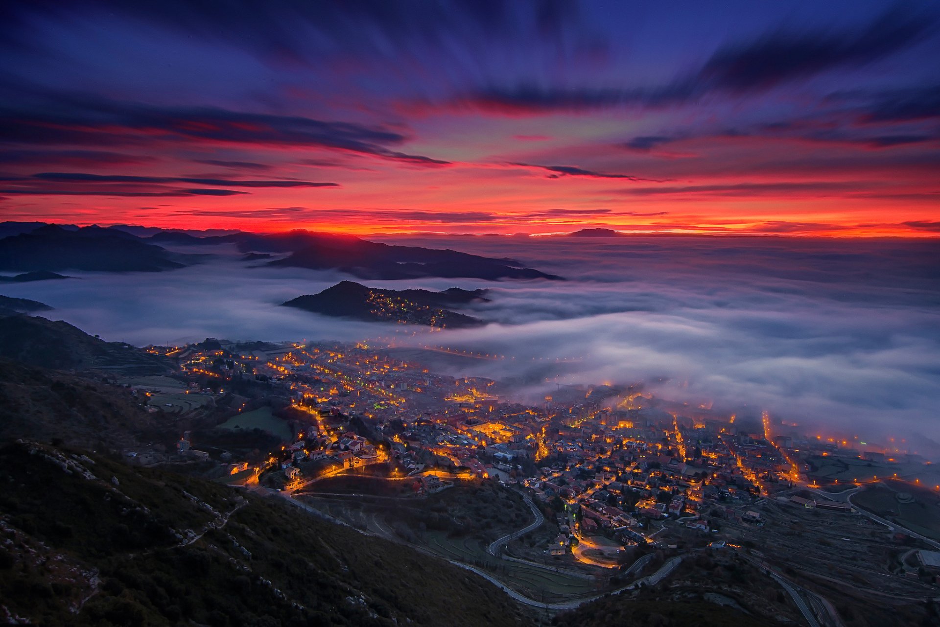 Night landscape photography of Berga, Catalonia, Spain, showing a fog-filled valley beneath a vibrant sky at the horizon with clouds and city lights glowing below.