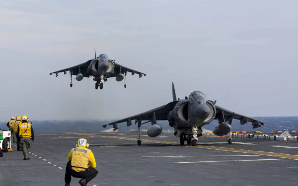 Two McDonnell Douglas AV-8B Harrier II jet fighter warplanes on an aircraft carrier deck, with crew members guiding the landing operation.