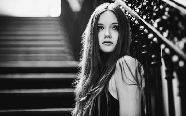 Black and white HD wallpaper of a model with long hair on a staircase, captured with a bokeh effect. The woman is by an intricately designed railing.