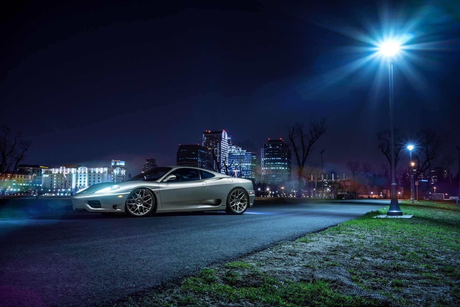 A sleek silver Ferrari 360 Modena supercar parked on an urban road at night with city lights and modern buildings in the background, captured in HD for a desktop wallpaper.
