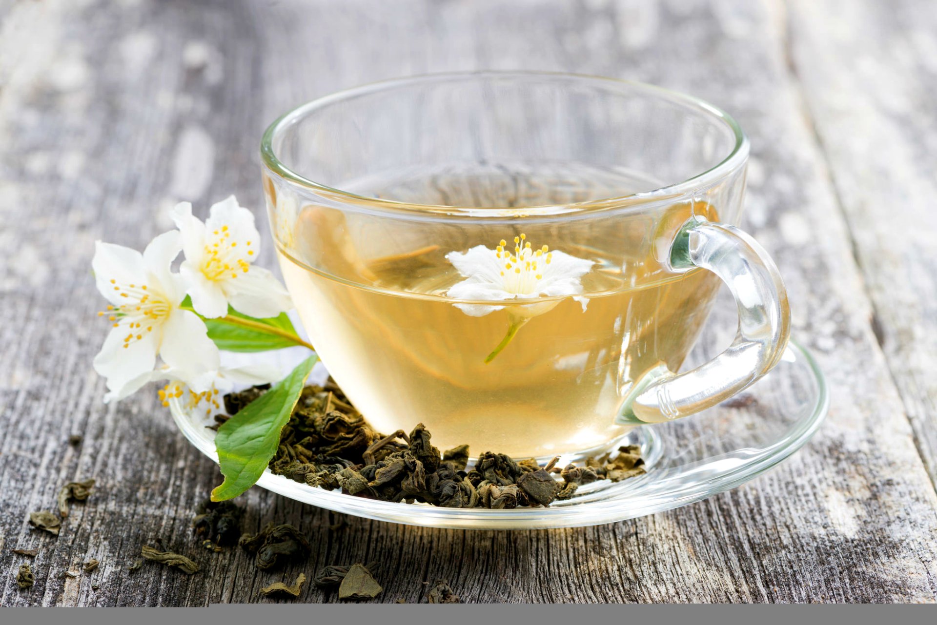HD PC desktop wallpaper showing a clear glass cup of jasmine tea with a jasmine blossom and loose tea leaves on a weathered wooden surface.