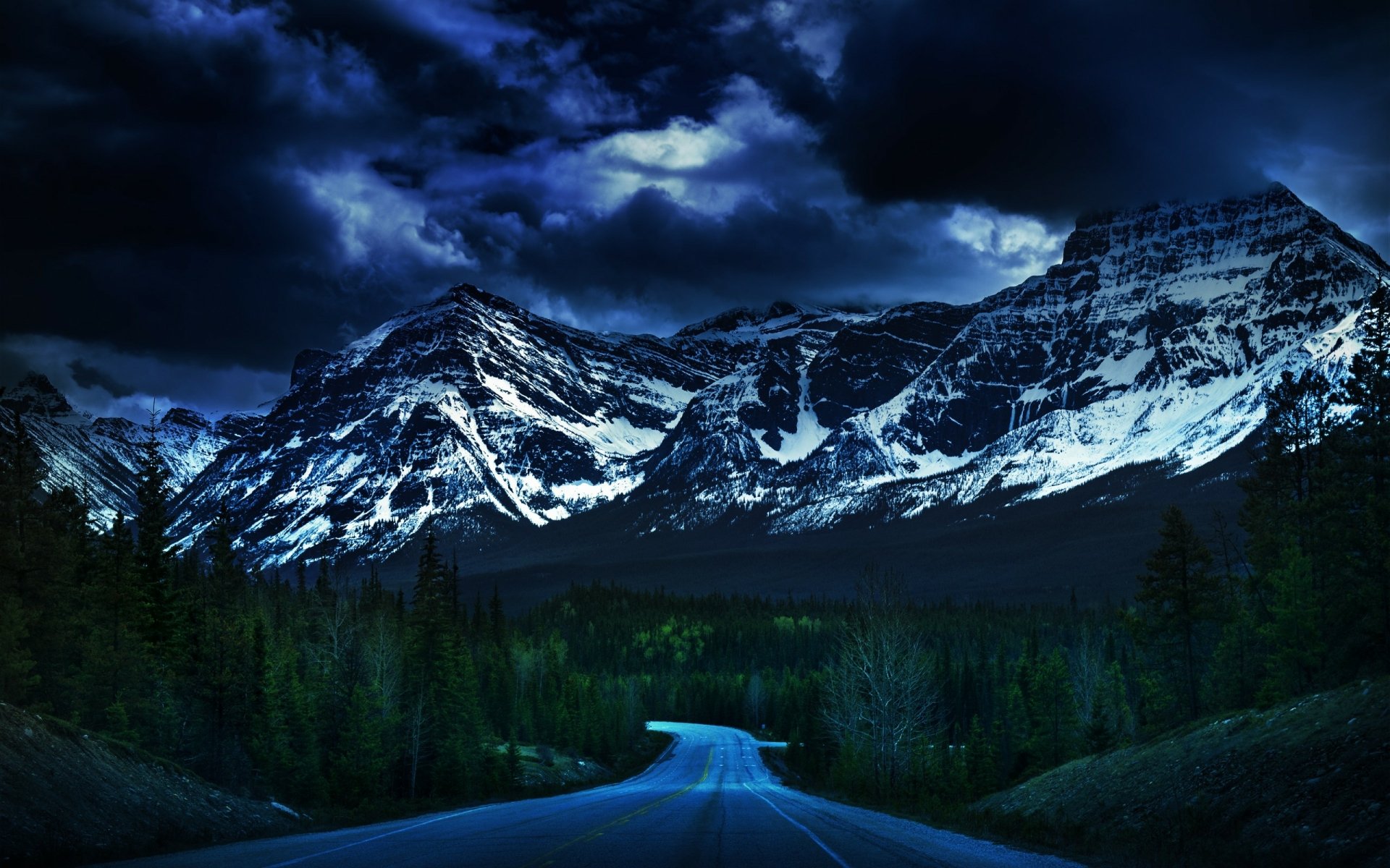 A dark, moody HD desktop wallpaper featuring a winding man-made road through a forest with towering snow-capped mountains under a cloudy night sky.