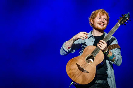English singer Ed Sheeran holding a guitar and smiling on stage under blue lighting. This HD desktop wallpaper captures his joyful performance moment.
