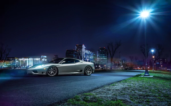 A sleek silver Ferrari 360 Modena supercar parked on an urban road at night with city lights and modern buildings in the background, captured in HD for a desktop wallpaper.