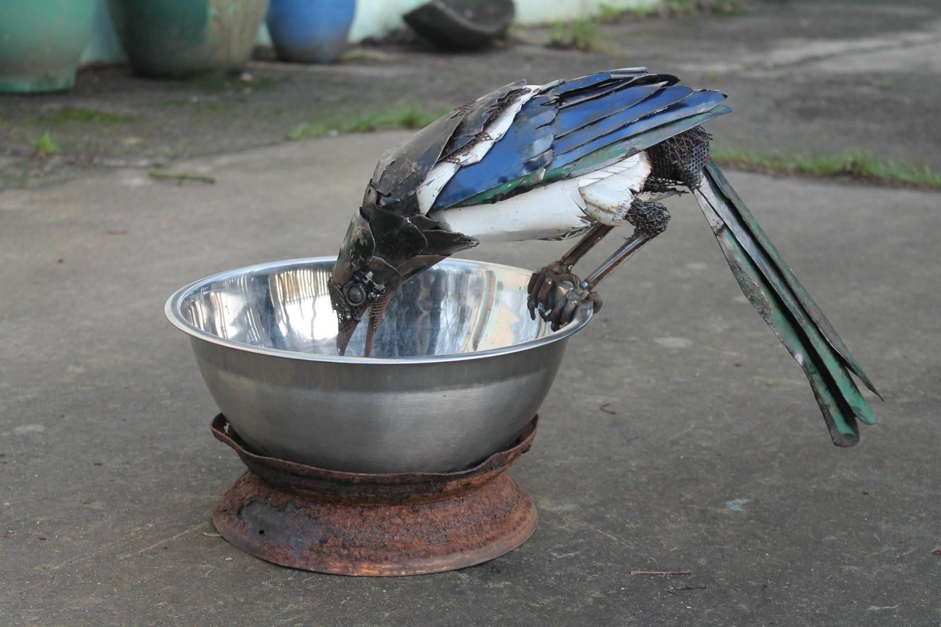 Scrap-metal bird sculpture drinking from a metal bowl on concrete — man-made artwork, HD PC desktop wallpaper and background.