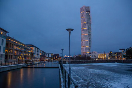 Night view of Turning Torso skyscraper in Malmö, Sweden, towering above waterfront buildings and an icy canal — man-made modern tower captured as a 5K Ultra HD PC desktop wallpaper.