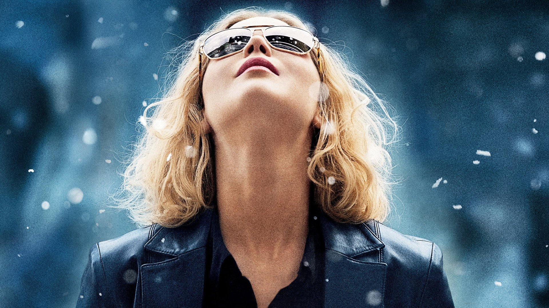 HD PC desktop wallpaper: blonde woman in sunglasses and leather jacket gazes upward amid falling snow — promotional-style portrait referencing the film Joy (2015).