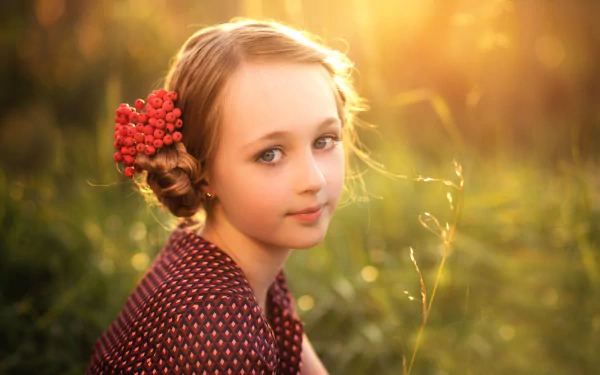 A serene child with blue eyes and blonde hair sits in a sunlit field, adorned with a floral accessory. The soft bokeh enhances the warm, dreamy atmosphere of the HD wallpaper.