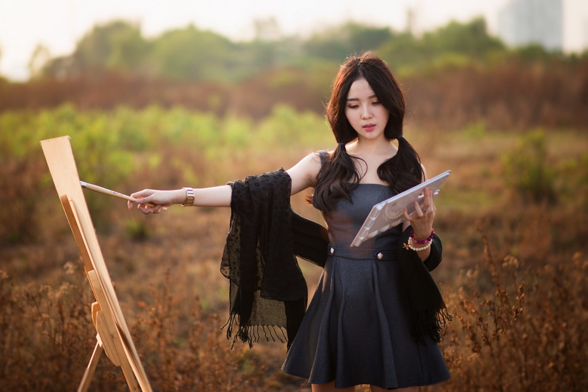 A brunette woman with an oriental look stands outdoors, painting on a canvas with a bokeh background. She holds a palette, emanating a calm mood. HD wallpaper and background.