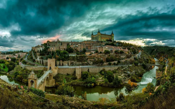 A panoramic view of Toledo, Spain, showcasing a blend of historic buildings, a bridge over a river, green trees, and mountains under a dramatic sky.