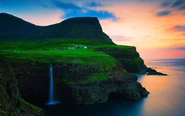 A stunning view of a waterfall cascading down a cliff in Gásadalur, Faroe Islands, surrounded by lush green landscapes and a picturesque village beneath a colorful sunset sky.
