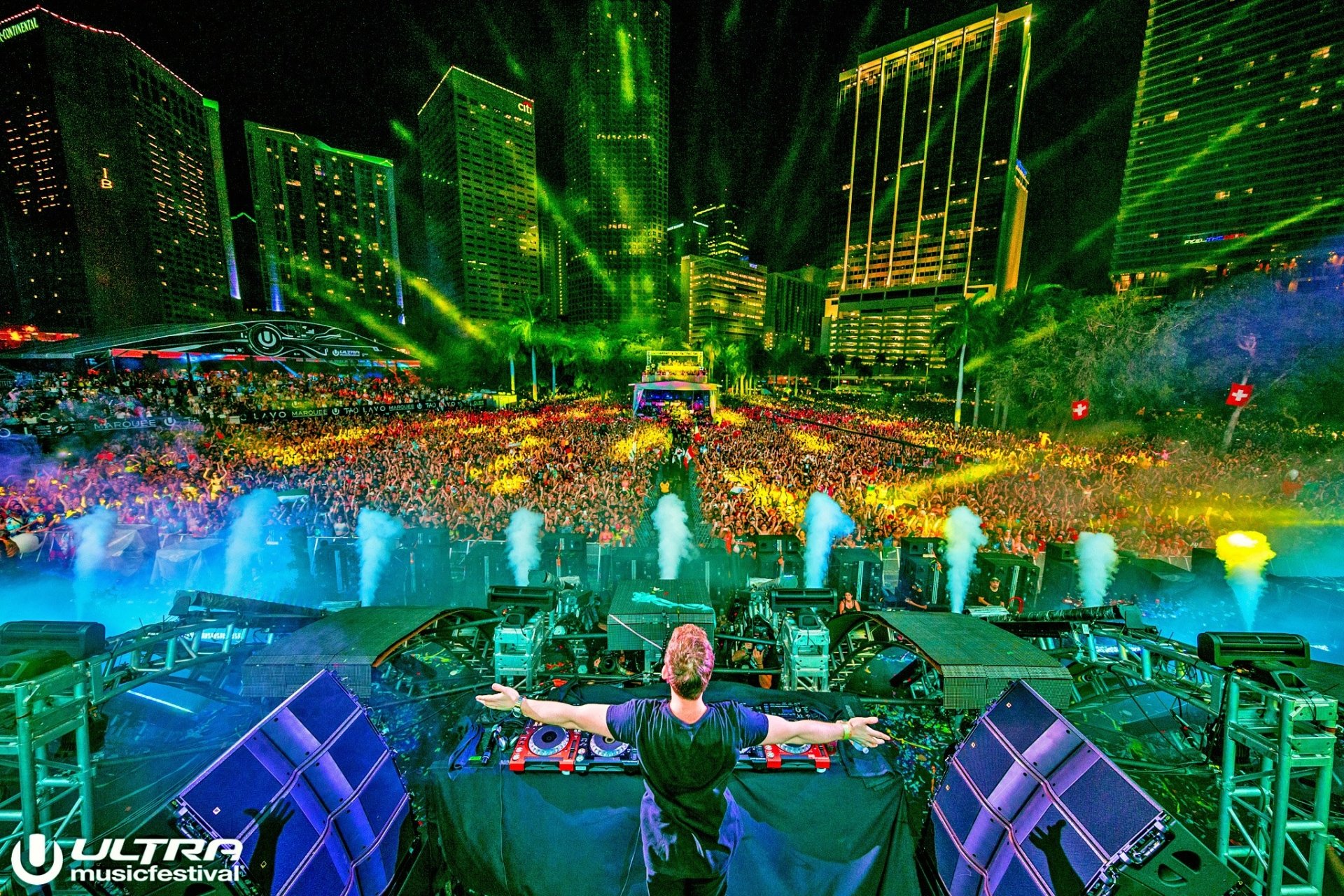 HD PC desktop wallpaper: music-themed festival scene — an EDM DJ on stage with outstretched arms before a neon-lit, packed crowd and glowing city skyline.