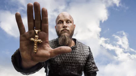 Ragnar Lothbrok from the TV show Vikings holds a golden crucifix, standing against a dramatic sky, creating a striking HD wallpaper for fans of the series.
