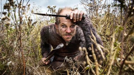 Floki, portrayed by Gustaf Skarsgård in the TV show Vikings (2013), is depicted in a rugged, overgrown landscape, wielding a weapon and showing a determined expression.