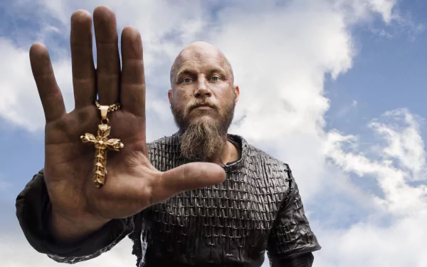 Ragnar Lothbrok from the TV show Vikings holds a golden crucifix, standing against a dramatic sky, creating a striking HD wallpaper for fans of the series.