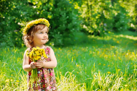A little girl in a floral dress smiles while holding a bouquet of dandelions, wearing a matching wreath, set against a vibrant summer backdrop of green grass and trees.