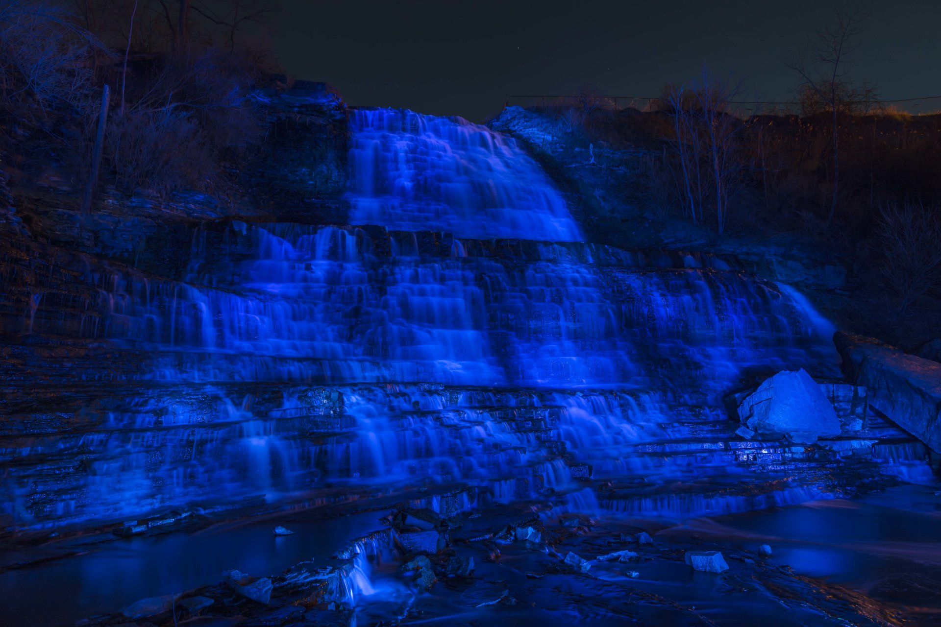 Albion Falls in Canada illuminated in blue at night, cascading over layered rocks; a serene 4K Ultra HD nature wallpaper showcasing the waterfall against a dark sky.